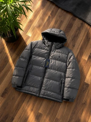 Mörkgrå pufferjacka med huva - Snygg mörkgrå pufferjacka från Polo Ralph Lauren med huva och dragkedja framtill. Jackan har två sidofickor med dragkedja och justerbar huva med snörning. Klassisk logga broderad på bröstet. Perfekt för kalla vinterdagar. Huva kan tas av med dragkedja. Kom dm om du har frågor eller är intresserad.