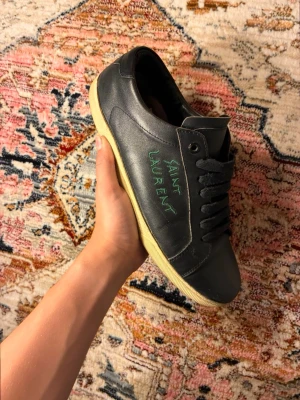 Svarta Saint Laurent sneakers - Svarta sneakers från Saint Laurent i läder med klassisk låg profil och svarta skosnören. Sidan pryds av grön text med märkesnamnet. Yttersulan är beige och ger en snygg kontrast till den svarta ovandelen. Perfekta för en stilren och trendig look. Patched i hälen inget som stör andvänding 