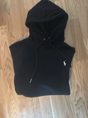 Svart hoodie från Polo Ralph Lauren - Klassisk svart hoodie från Polo Ralph Lauren med vit broderad logga på bröstet. Hoodien har justerbar dragsko i huvan och en stor magficka. Perfekt oversized fit och mjukt material som känns riktigt skönt mot huden.