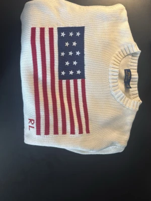 Vit stickad Ralph lauren tröja med USA-flagga - Vit stickad tröja med rund hals och ribbade muddar. På bröstet finns en stor USA-flagga i blått, rött och vitt samt broderade bokstäver 'RL'. Tröjan har en klassisk passform och är perfekt för dig som gillar preppy stil. Passar som S/M