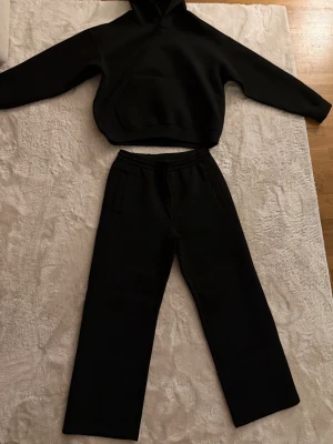 Svart scubaset med hoodie och byxor - Säljer ett svart loungewear-set med hoodie och matchande byxor. Hoodien har en stor ficka framtill och byxorna har raka ben och resår i midjan. Perfekt för chill dagar hemma eller när du vill vara extra bekväm.