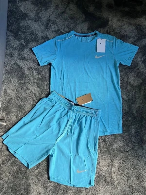Nike ljusblått träningsset - Säljer ett ljusblått träningsset från Nike med t-shirt och shorts. Setet har en sportig look med Nike-logga på både tröjan och shortsen. Materialet är lätt och andas, perfekt för träning eller löpning. Shortsen har resår i midjan och dragsko.