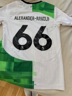 Signerad Alexander-Arnold fotbollströja - Säljer en vit och grön Nike fotbollströja med Alexander-Arnold och nummer 66 på ryggen. Tröjan har svarta detaljer, Premier League-logga och är tillverkad i Dri-FIT-material för bra ventilation. Perfekt för dig som älskar fotboll och vill sticka ut på planen.