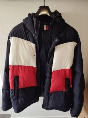 Färgglad pufferjacka med huva - Pufferjacka med cool färgblock-design i svart, vitt och rött. Jackan har huva, dragkedja och två fickor framtill. Quiltad look och oversized passform som håller dig varm under vintern.