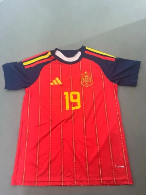 Spanien tröja med Lamine Yamal 19 - Säljer en röd matchtröja från Spaniens landslag med gula detaljer och nummer 19 på bröstet. Tröjan har mörkblå ärmar, gula ränder på axlarna och Adidas-logga. Tillverkad i lätt funktionsmaterial, perfekt för fotbollsträning eller match. Storlek L Helt ny utan prislapp 
