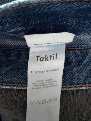 Blå raka jeans från Taktil - Säljer ett par blå jeans från Taktil i modellen T Herman Straight. Jeansen har raka ben, klassisk femficksdesign och råa, fransiga benslut för en avslappnad look. Perfekta till en streetwear-outfit eller för dig som gillar enkel denimstil. Jeansen är cropped hemma alltså inte tillverkade med raw hem. Storleken är inte helt säker men skulle säga sitter ungefär som 29/30-30/30 nypris runt 500-700 tror jag. Har ett litet hål vid en av bakfickorna.