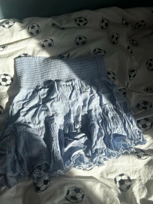 Ljusblå volangkjol med smock - Supersöt ljusblå kortkjol med bred smock i midjan och volangdetaljer. Kjolen har ett diskret småprickigt mönster i rosa och är gjord i ett mjukt bomullstyg. Perfekt för dig som gillar en feminin och avslappnad stil.