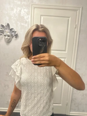 Vit blus med volangärmar - Säljer en söt vit blus med volangdetaljer på ärmarna och ett diskret mönster i tyget. Blusen har rund halsringning och är tillverkad i ett mjukt material som känns luftigt och skönt. Perfekt för dig som vill ha en stilren men ändå unik topp.