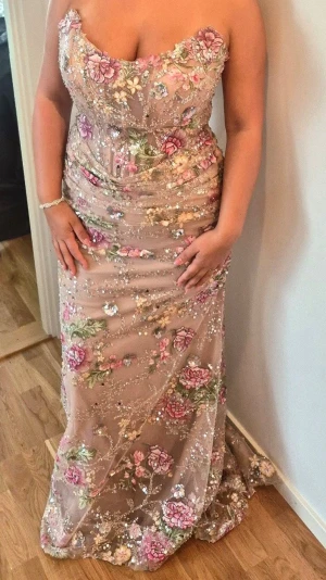 Beige långklänning med blommor och paljetter - Supersnygg beige långklänning med figurnära passform, täckt av rosa och gröna blommor samt glittriga paljetter. Klänningen har en elegant släp och transparent mesh med broderade detaljer. Perfekt för bal eller festkväll! Armar ingår! Jag är 165 lång och bär medium/large vanligtvis. 