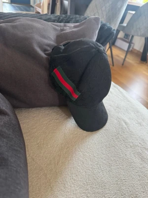 Svart keps med grön-röd rand - Säljer en svart keps med diskret mönster och en grön-röd rand på sidan. Kepsen har böjd skärm och justerbart spänne bak. Materialet ser ut att vara bomull eller canvas och designen är riktigt clean och stilren.
