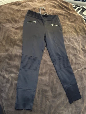 Svarta leggings med dragkedjor - Snygga svarta leggings med silverfärgade dragkedjor framtill och vid bensluten. De har ribbade detaljer över knäna och en tight passform. Perfekta för en trendig och bekväm look. Materialet ser stretchigt ut och ger en cool biker-vibe.