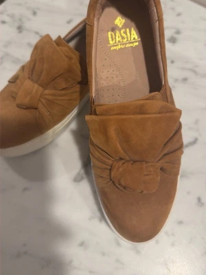 Bruna loafers i mocka från Dasia - Säljer ett par snygga bruna loafers från Dasia i mjuk mocka. Skorna har en chunky vit sula och en stor dekorativ knut framtill som ger en cool och trendig look. Perfekta för dig som vill ha både stil och komfort i vardagen.