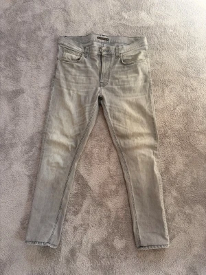 Nudie jeans lean dean - Säljer ett par ljusgrå slim fit jeans med klassisk femficksdesign och snygga sömmar på bakfickorna. Jeansen har dragkedjegylf och raka ben. Perfekta för dig som gillar en clean och stilren look. Storleken är 31/34 men är uppsydda till 32 i längd. Tveka inte med att be om fler bilder och går ner i pris vid snabb affär. 