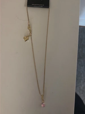 Guldhalsband med rosa pärla - Snyggt och tunt guldhalsband med en liten rosa pärla som hänge. Kedjan har ett hjärtformat detaljerat lås och en enkel, elegant design. Perfekt för dig som gillar stilrena accessoarer med en söt touch.