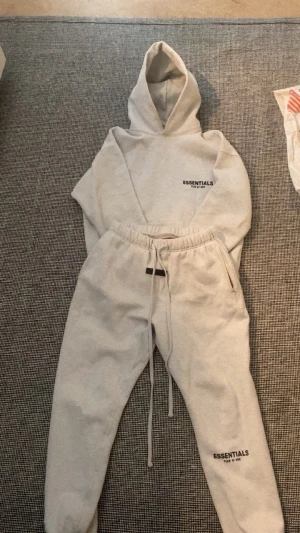 Grå Essentials tracksuit - Ljusgrå tracksuit från Essentials med huva och tryckt logga på bröstet. Tracksuit har en avslappnad passform och ribbade muddar vid ärmslut och nederkant. Materialet är mjukt och passar perfekt för en streetwear-look.