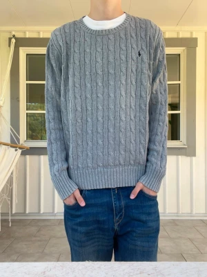 Grå kabelstickad tröja från Ralph Lauren - Snygg grå kabelstickad tröja med rund halsringning och ribbade muddar vid ärmslut och nederkant. Tröjan har ett diskret broderat märke på bröstet och är perfekt för lager-på-lager. Passar till jeans eller chinos för en avslappnad look.
