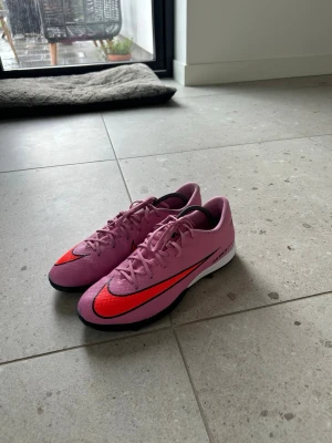 Rosa Nike Mercurial fotbollsskor - Säljer ett par rosa Nike Mercurial fotbollsskor med röd och svart swoosh på sidan. Skorna har låg profil, snörning och en slitstark yttersula för bra grepp på planen.