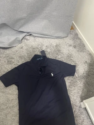  Polo Ralph Lauren, PRIS GÅT ATT DISKUTERA  - Klassisk mörkblå pikétröja från Polo Ralph Lauren med vit broderad logga på bröstet. Tröjan har korta ärmar, krage och knappar framtill. Perfekt för en stilren och avslappnad look.