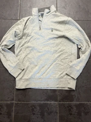 Ljusgrå halvzip sweatshirt - Ljusgrå sweatshirt med halv dragkedja och ståkrage. Tröjan har långa ärmar, ribbade muddar och en liten broderad logga på bröstet. Perfekt för en avslappnad och stilren look. Storlek M, helt ny aldrig använd.