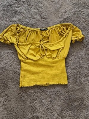 Gul croppad topp från Boohoo - Trendig gul croppad topp från Boohoo med offshoulder-design och korta puffärmar. Toppen har smockad midja och en söt knytning framtill.