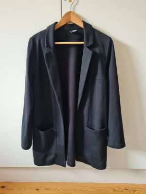 Svart lång kavaj med fickor från H&M strl S - Stilren svart kavaj med två stora framfickor. Klassisk krage och enkel design utan synliga knappar. Material 56% akryl, 33% polyester, 9% bomull, 2% elastan. Storlek S.