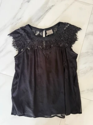 Svart blus med spetsdetaljer Vero Moda - Snygg svart blus från Vero Moda med spetsdetaljer över axlar och bröst. Blusen är ärmlös och har en rund halsringning samt knapp i nacken. Storlek M men passar mer S!! 
