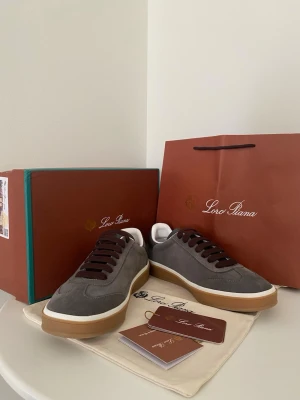 Grå mockasneakers från Loro Piana - Stilrena sneakers i grå mocka från Loro Piana med bruna skosnören och vit/brun sula. Låg modell med klassisk siluett och diskreta detaljer. Perfekta för dig som gillar exklusiv och tidlös design. Kommer med originalpåse och förpackning.