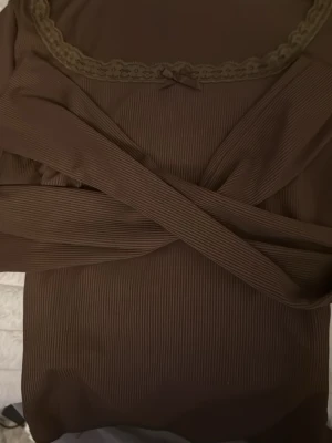 Brun ribbad topp med spetsdetalj - Säljer en brun ribbad topp med lång ärm. Toppen har en söt spetskant och liten rosett vid halsringningen. Perfekt för dig som gillar enkla men fina detaljer. Passar till många olika stilar.