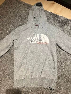 Grå hoodie från The North Face - Snygg grå hoodie från The North Face med stor logga i vitt framtill. Klassisk känguruficka, justerbar huva med dragsko och mjukt, borstat innerfoder. Perfekt för chill dagar eller när du vill ha en clean streetstyle-look.