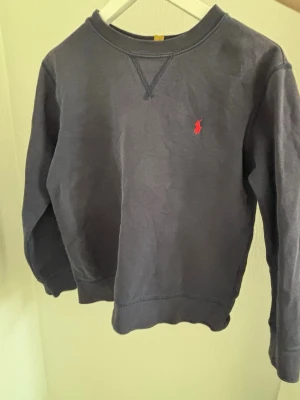 Mörkblå sweatshirt från Polo Ralph Lauren - Klassisk mörkblå sweatshirt från Polo Ralph Lauren med rund halsringning och röd broderad logga på bröstet. Tröjan har ribbade muddar vid ärmslut och nederkant samt en avslappnad passform. Perfekt för en chill och stilren look. Barnstorlek 