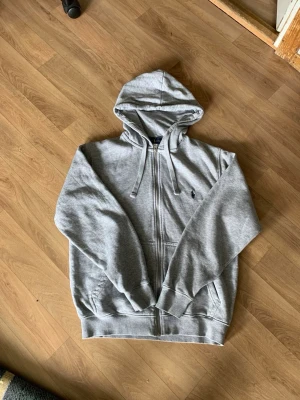 Grå  hoodie från Polo Ralph Lauren - Säljer en grå hoodie med dragkedja från Polo Ralph Lauren. Klassisk design med huva och dragsnören, två fickor fram och den ikoniska broderade loggan på bröstet. Tillverkad i mjukt sweatshirtmaterial som är riktigt skönt.