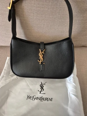 Svart axelremsväska  - Stilren svart axelremsväska med ikoniskt guldigt YSL-emblem framtill. Väskan har justerbar rem och minimalistisk design som ger en lyxig känsla. Perfekt för dig som gillar klassiska och trendiga accessoarer.
