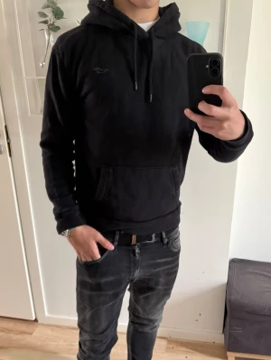  Hollister hoodie  - Svart hoodie från Hollister med klassisk huva och dragsnören. Tröjan har en stor magficka och diskret Hollister-logga på bröstet. Tillverkad i mjukt bomullsmaterial och har ribbade muddar vid ärmslut och nederkant. Perfekt för en avslappnad och stilren look.