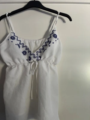 Vit linnetopp med blå broderi - Säljer en söt vit linnetopp med tunna axelband och blå broderade detaljer vid urringningen. Toppen har knyt i midjan och är lätt och luftig, perfekt för varma dagar. Passar dig som gillar en avslappnad och somrig stil.