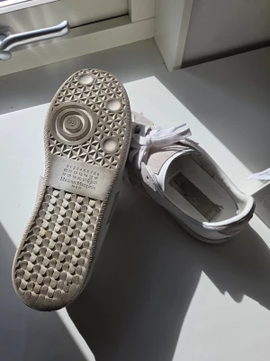 Maison Margiela vita sneakers - Maison Margiela sneakers i vitt med beige detaljer. Skorna har en klassisk låg silhuett, vita skosnören och en sula med unikt mönster och logotyp. Materialet är en mix av mocka och läder för en stilren och exklusiv look.