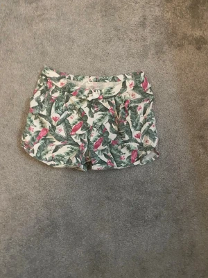 Gröna blommiga shorts med snörning - Säljer ett par somriga shorts med grönt och rosa blommönster. Shortsen har snörning i midjan och är tillverkade i ett lätt och mjukt material, perfekta för varma dagar. Passformen är loose och de har en avslappnad look.