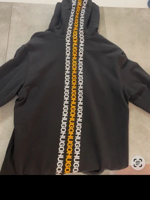 Svart hoodie med HUGO-logga - Storlek S men passar MSvart hoodie från HUGO med stor ficka framtill och huva med dragsko. På bröstet finns en gul HUGO-logga och längs ryggen går ett vitt och gult HUGO-mönster hela vägen från huvan ner. Perfekt för en chill och trendig look. 