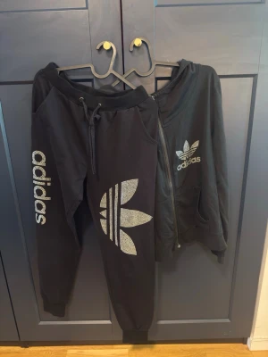 Svart Adidas mysdress med strassdetaljer - Säljer en svart Adidas mysdress med byxa och zip-hoodie. Båda plaggen har stora Adidas-loggor i strass och byxan har även Adidas-text längs benet. Mjuk bomullskänsla och avslappnad passform, perfekt för chill eller träning.