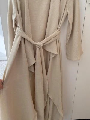 Beige morgonrock med knytskärp - Stilrent och elegant 2-delat set i beige från Shein. Består av en ärmlös maxiklänning/innerklänning och en matchande långärmad tunn cardigan/kimono med knytband i midjan. Tillverkat i ett lätt, luftigt material med fin struktur. Perfekt för en lager-på-lager-look eller blygsamt mode (modest fashion).