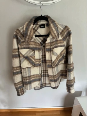 Beige och brun rutig overshirt - Säljer en snygg overshirt från Vero Moda i beige, brunt och blått rutigt mönster. Jackan har två stora bröstfickor med lock, knappar framtill och en mjuk, borstad yta. Perfekt för lager på lager och trendig look.