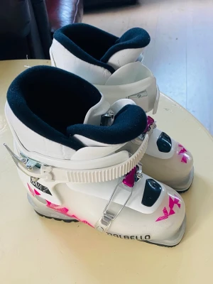 Vita Dalbello junior  pjäxor med rosa detaljer nyskick 22,5 - Säljer ett par vita Dalbello Gaia 2 skidpjäxor med rosa och svarta detaljer. Pjäxorna har två spännen för bra passform och svart innermaterial. Perfekta för dig som vill ha snygga och funktionella pjäxor i backen. 22,5 mostavae 35-36 i skor.