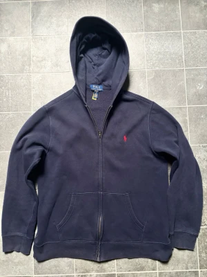 Mörkblå hoodie från Polo Ralph Lauren - Mörkblå hoodie med dragkedja från Polo Ralph Lauren. Klassisk design med röd broderad logga på bröstet, kängurufickor och huva. Tillverkad i mjuk bomullsblandning som känns skön mot huden. Perfekt för en avslappnad och stilren look.