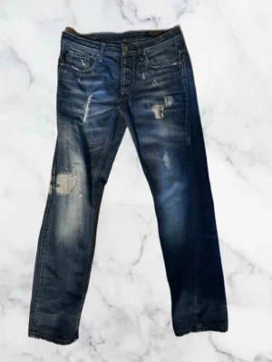 Jack and Jones jeans  - Säljer ett par blå jeans med slitningar och patchdetaljer på benen. Jeansen har klassisk femficksmodell, raka ben och en snygg tvättad look. Perfekta för dig som gillar en avslappnad och trendig stil. W30 L32