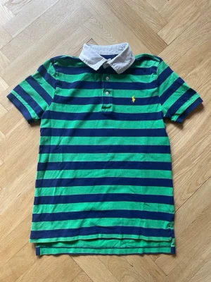 Piké Polo Ralph Lauren - Snygg pikétröja från Polo Ralph Lauren med gröna och marinblå ränder, vit krage och klassisk gul logga på bröstet säljes. Tröjan är i storlek 10-12 år och i mycket fint skick. 