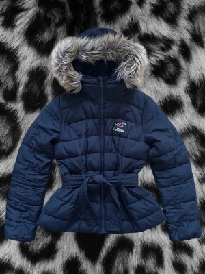 Hollister jacka  - Asfet mörkblå pufferjacka från Hollister med huva och fuskpälsdetalj. Jackan har bälte i midjan för en schysst siluett och dragkedja framtill samt rosa innerfoder. Perfekt för kalla dagar och riktigt varm. Klassisk Hollister-logga på bröstet. Jackan är köpt 2012 så finns ej att köpa idag. Storlek M. Alla mått finns med på bilderna.