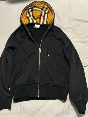 burberry kofta - Säljer en svart hoodie med dragkedja och kängurufickor. Insidan av huvan har ett klassiskt rutigt mönster i beige, svart, rött och vitt. Tröjan har ribbade muddar vid ärmslut och nederkant samt justerbara snören i huvan. Perfekt för dig som gillar stilrena detaljer.