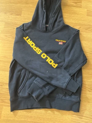 Mörkblå hoodie Polo Sport - Mörkblå hoodie från Polo Sport med gul logga på ärmen och liten broderad text samt amerikansk flagga på bröstet. Klassisk huva och känguruficka framtill. Perfekt för en sportig och avslappnad stil.