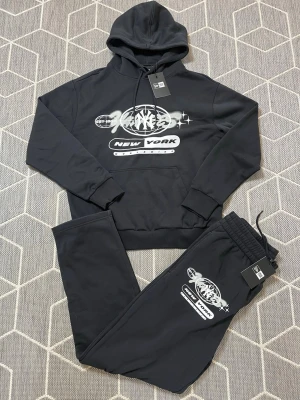 New Era Tracksuit - Snyggt svart loungewear set med hoodie och matchande byxor. Båda plaggen har ett stort grafiskt New Era-tryck i vitt och grått framtill. Hoodien har huva med snörning och magficka, byxorna har resår i midjan och tryck på benet. Perfekt för chill dagar hemma.