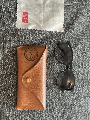 Svarta Ray-Ban solglasögon - Snygga svarta solglasögon från Ray-Ban med klassisk wayfarer-form. Bågarna är i svart plast och glasen är mörka. Medföljer originalfodral i brunt läder och putsduk med Ray-Ban-logga. Perfekta för en cool och tidlös stil.