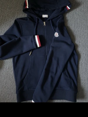 Mörkblå hoodie med dragkedja och huva - Mörkblå hoodie från Moncler med dragkedja, fickor fram och huva. Detaljer i rött, vitt och blått på huva och ärmslut samt Moncler-logga på bröstet. Tillverkad i mjukt material som känns skönt mot huden. Perfekt för en avslappnad och stilren look.